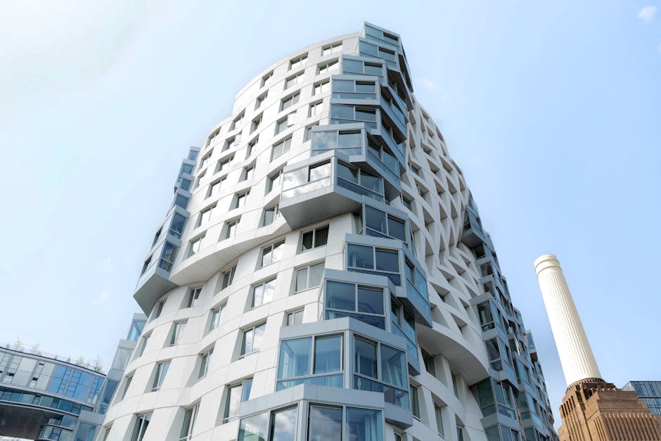 A modern, high-rise residential building with a distinctive curved facade featuring multiple protruding glass balconies, set against a clear blue sky. The building has a predominantly white exterior w