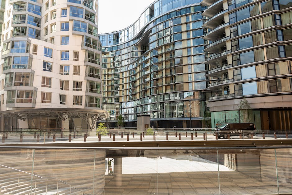 Exterior view of a modern residential building with curved glass and white panels, featuring multiple floors with large windows and balconies. The scene shows a paved area in front of the building with a row of short wooden and metal posts along the pavement edge. A black van is parked near one side of the building, partially visible behind glass barriers. The environment appears clean and well-maintained, with natural daylight illuminating the scene. This setting is relevant to house removals and furniture transport, indicating the logistics involved in home relocation through professional services like Battersea Man and Van, who handle packing, loading, and moving within urban apartment buildings.