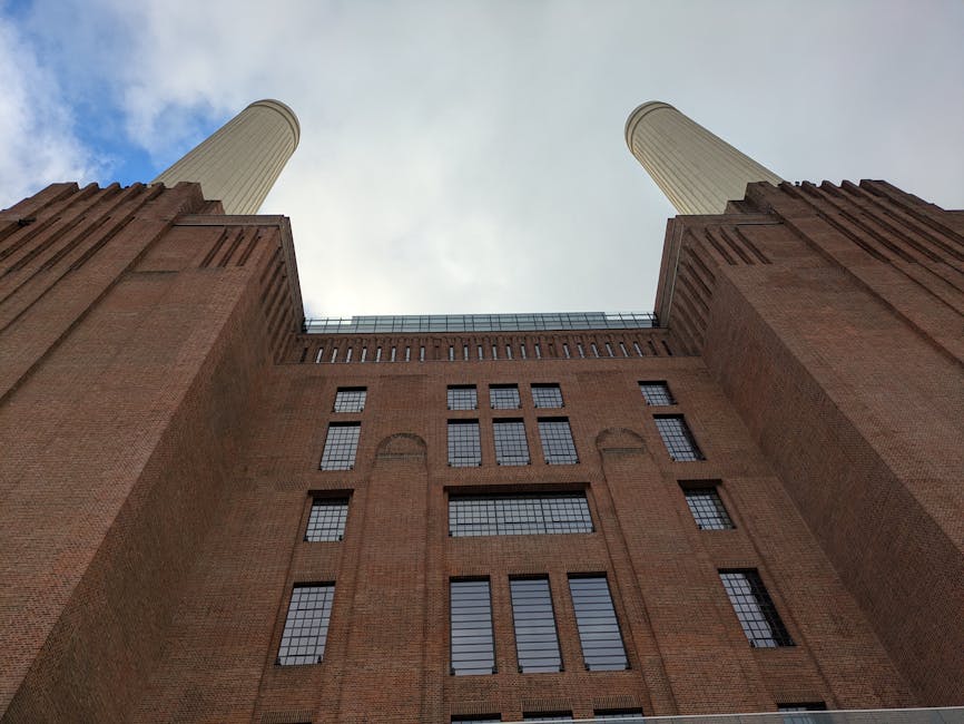 A low-angle view of a large brick building with multiple rectangular windows and two prominent cylindrical chimneys extending upwards against a cloudy sky. The building's façade features brickwork in reddish-brown tones with vertical and horizontal architectural details. The windows vary in size, some with grid-like panes, and are set within arched and rectangular frames. The image captures the exterior of a historic or industrial structure, possibly part of a factory or power station, with the perspective emphasizing the building's height and imposing presence. This setting illustrates the type of building that might be involved in house removals or relocations when moving from or to such industrial-style premises, with Battersea Man and Van providing specialist moving services for properties in similar environments. The scene is illuminated by natural daylight, highlighting the textures of the brick and metal elements.