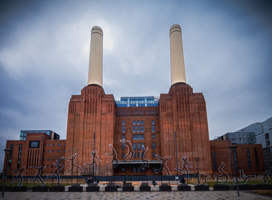 A large red-brick building with two tall, white chimneys on either side, situated at Battersea Power Station in London. The building features a central entrance with steps leading up to it, and several small windows arranged symmetrically along the facade. In front of the building, there are decorative metal sculptures of human figures performing various action poses, mounted on black rectangular bases. The sculptures are illuminated by subtle lighting, and the pavement in the foreground is made of patterned stone tiles. The sky above is overcast with dark clouds, contributing to a moody atmosphere. This scene is part of a cityscape reflecting the urban environment around Battersea Power Station, which is of relevance to moving and relocation services such as those provided by Battersea Man and Van, especially in the context of home relocation or furniture transport within the area.